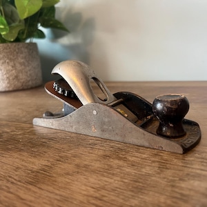 Puede incluir: Un cepillo de mano vintage de metal con mango de madera. El cepillo tiene un acabado plateado y una perilla negra. El cepillo está sobre una superficie de madera.