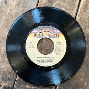 May include: A black vinyl record with a colorful label featuring the text "Casablanca Records and Filmworks" and the song title "Maniac (Instrumental)" by Michael Sembello. The record is on a wooden surface.