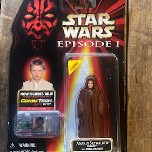 Könnte beinhalten: Star Wars Episode I Actionfigur von Anakin Skywalker in einem braunen Gewand, verpackt mit einer Comlink-Einheit. Die Verpackung zeigt das Gesicht von Darth Maul, das Star Wars-Logo und den Text "NOW FIGURES TALK!". Alter 4+