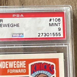May include: A basketball trading card encased in a clear plastic holder. The card features the Knicks logo and the name of the player, Kiki Vandeweghe. The card is graded by PSA, with a grade of 9 and a serial number 27301555.