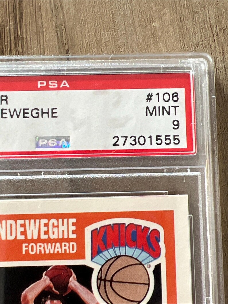 May include: A basketball trading card encased in a clear plastic holder. The card features the Knicks logo and the name of the player, Kiki Vandeweghe. The card is graded by PSA, with a grade of 9 and a serial number 27301555.