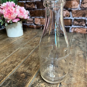 May include: A clear glass milk bottle with a textured surface. The bottle has a narrow neck and a rounded bottom. The bottle is empty and sitting on a wooden surface.