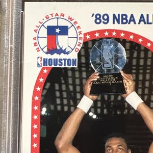 Pu&ograve; includere: Un giocatore di basket tiene in alto un trofeo con la scritta 'Most Valuable Player All-Star Game'. Il trofeo &egrave; una palla da basket di vetro trasparente con una base argentata. Il giocatore indossa una maglia bianca con un logo rosso, bianco e blu sul davanti. Il logo presenta una stella e la parola 'Houston'. Lo sfondo &egrave; un'arena buia con una folla di persone sugli spalti. Il testo '89 NBA All-Star Weekend' &egrave; in alto nell'immagine.