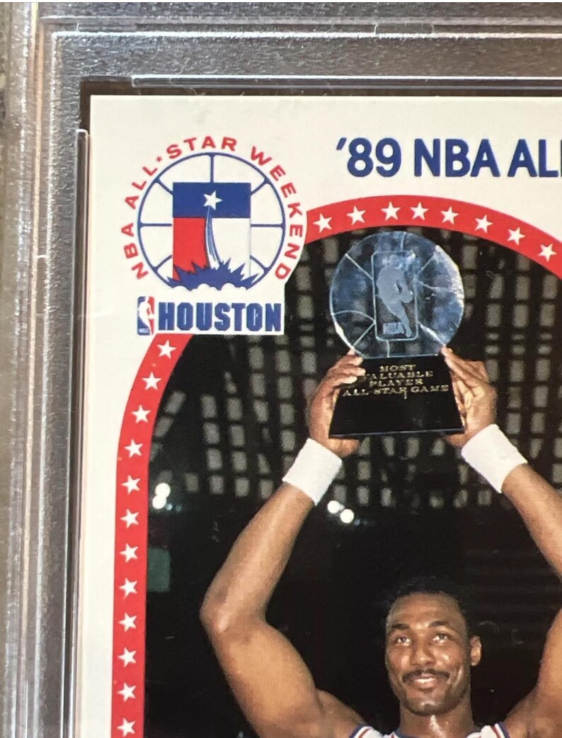 Pu&ograve; includere: Un giocatore di basket tiene in alto un trofeo con la scritta 'Most Valuable Player All-Star Game'. Il trofeo &egrave; una palla da basket di vetro trasparente con una base argentata. Il giocatore indossa una maglia bianca con un logo rosso, bianco e blu sul davanti. Il logo presenta una stella e la parola 'Houston'. Lo sfondo &egrave; un'arena buia con una folla di persone sugli spalti. Il testo '89 NBA All-Star Weekend' &egrave; in alto nell'immagine.