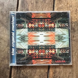 May include: A CD case featuring the album "Vegas" by The Crystal Method. The cover art displays a symmetrical, abstract design with red, orange, and gray hues. The text "the crystal method" is vertically aligned on the left side of the case.