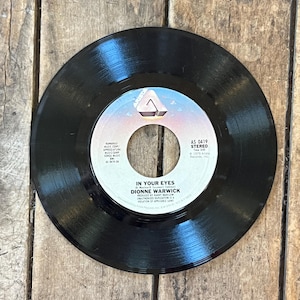 Puede incluir: Un disco de vinilo negro con una etiqueta blanca y azul. La etiqueta presenta el texto "IN YOUR EYES" de Dionne Warwick. El disco está centrado en una superficie de madera, mostrando su diseño clásico.