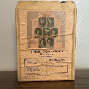 May include: A vintage Three Dog Night "Harmony" 8-track tape. The album cover features a sepia-toned image of the band members. The track list includes songs like "You" and "Never Been to Spain". The tape is in a worn, off-white plastic case.