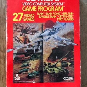 Peut inclure: Une boîte de cartouche de jeu Atari rouge avec une image d'un avion de chasse et de chars. La boîte indique "COMBAT" et "27 VIDEO GAMES". Le jeu comprend Tank, Tank Pong, Biplane, Invisible Tank, Jet Fighter et Deux Joueurs.