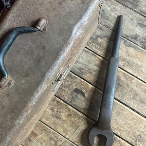 Könnte beinhalten: Eine rostige Metallwerkzeugkiste mit einem schwarzen Griff und einem großen, schwarzen Schraubenschlüssel mit U-förmigem Kopf.