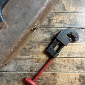 Peut inclure: Un coupe-tube vintage rouge et noir avec une poignée en métal et une poignée en plastique rouge. Le coupe-tube est posé sur une surface en bois.