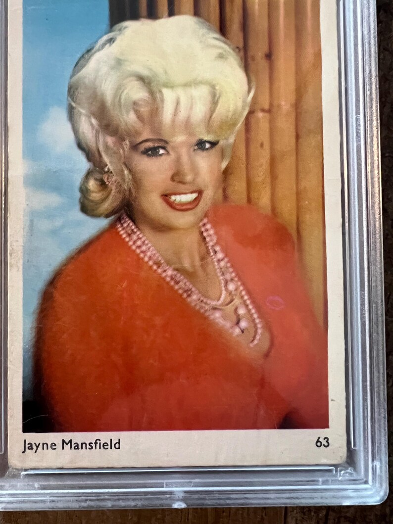 May include: A vintage trading card featuring a portrait of Jayne Mansfield. She is wearing a red sweater and a pearl necklace. The card has a white background with the text "Jayne Mansfield" and "63" printed on it.