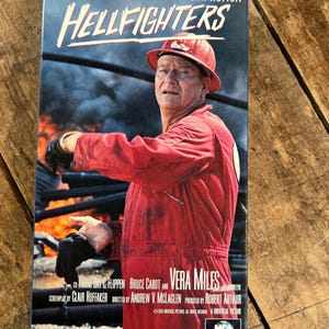 May include: A VHS tape for the movie "Hellfighters" featuring John Wayne. The cover shows Wayne in a red jumpsuit and hard hat, with the film's title in white. The background depicts a fiery scene.