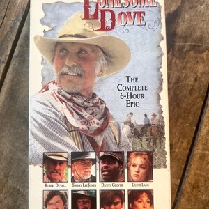 Pode incluir: Uma capa de fita VHS para o filme Lonesome Dove. A capa apresenta um close-up de Robert Duvall como Augustus McCrae, um caubói usando um chapéu branco e uma bandana vermelha. O texto na capa diz "Lonesome Dove", "The Complete 6-Hour Epic", e lista os membros do elenco, incluindo Robert Duvall, Tommy Lee Jones, Danny Glover, Diane Lane, Robert Urich, Frederic Forrest, Rick Schroder e Anjelica Huston. A capa também inclui o logotipo da Cabin Fever Entertainment Inc.