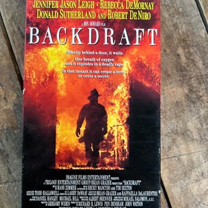 Può includere: Una videocassetta VHS del film "Backdraft" con Kurt Russell. La copertina mostra un pompiere in silhouette contro uno sfondo di fiamme. Il titolo "BACKDRAFT" è in grandi lettere, con i nomi degli attori sopra.