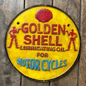 May include: A yellow round cast iron sign with black lettering that reads "Golden Shell Lubricating Oil for Motorcycles". The sign features a red Shell logo and two black figures on either side of the text.