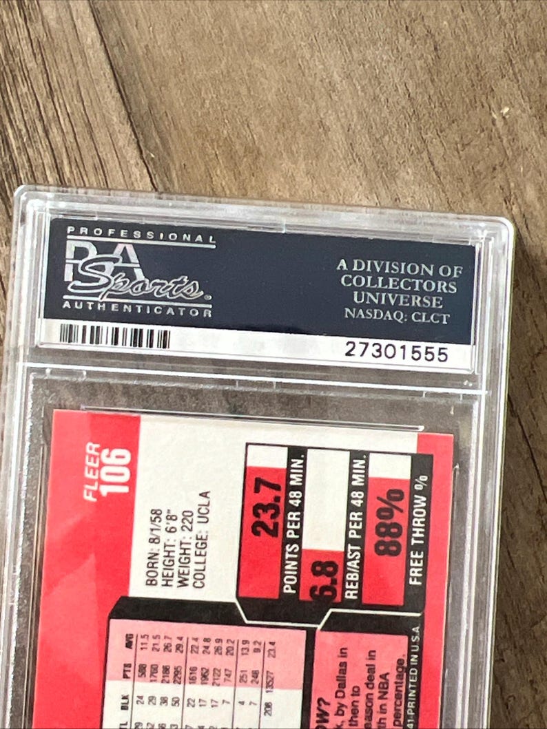 May include: A graded sports trading card in a protective case. The card features a red and white design with player statistics, including points per 48 minutes and free throw percentage. The case is labeled "PSA Sports Authenticator."