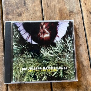 May include: A CD case with the album "Become What You Are" by The Juliana Hatfield Three. The cover art depicts a person's head and shoulders against a backdrop of green grass. The text "the JULIANA HATFIELD three" and "BECOME WHAT YOU ARE" are visible.