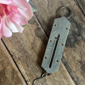 Puede incluir: Una balanza colgante vintage de plata con una aguja negra y una cara blanca. La balanza tiene marcas para onzas y gramos. La balanza está colgada de un gancho de metal.