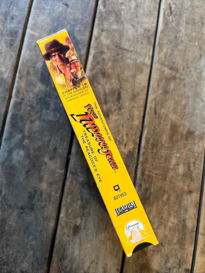 May include: A yellow VHS tape box for "The Adventures of Young Indiana Jones: Treasure of the Peacock's Eye." The box features images of Indiana Jones and the text "Chapter 18" and the Lucasfilm and Paramount logos.