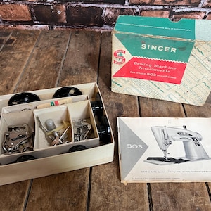 May include: Vintage Singer sewing machine attachments and accessories, including bobbins, presser feet, and a manual. The box is beige with black wheels and compartments. The box has a green, red, and white Singer box.