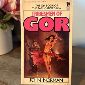 May include: A vintage paperback book titled "Tribesmen of Gor" by John Norman. The cover features a woman in a revealing outfit, with the title in large pink letters. The book is propped up on a small wooden easel.