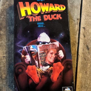 May include: A vintage VHS tape for the movie "Howard the Duck." The cover features the title in yellow and red, with a cartoon duck reading a magazine. The magazine cover features a woman with the text "Rolling Egg".