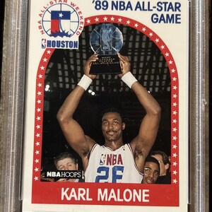 Pu&ograve; includere: Una carta da collezione del NBA All-Star Game con Karl Malone che tiene un trofeo. La carta ha un bordo bianco con uno sfondo rosso e il testo '89 NBA All-Star Game' in bianco.