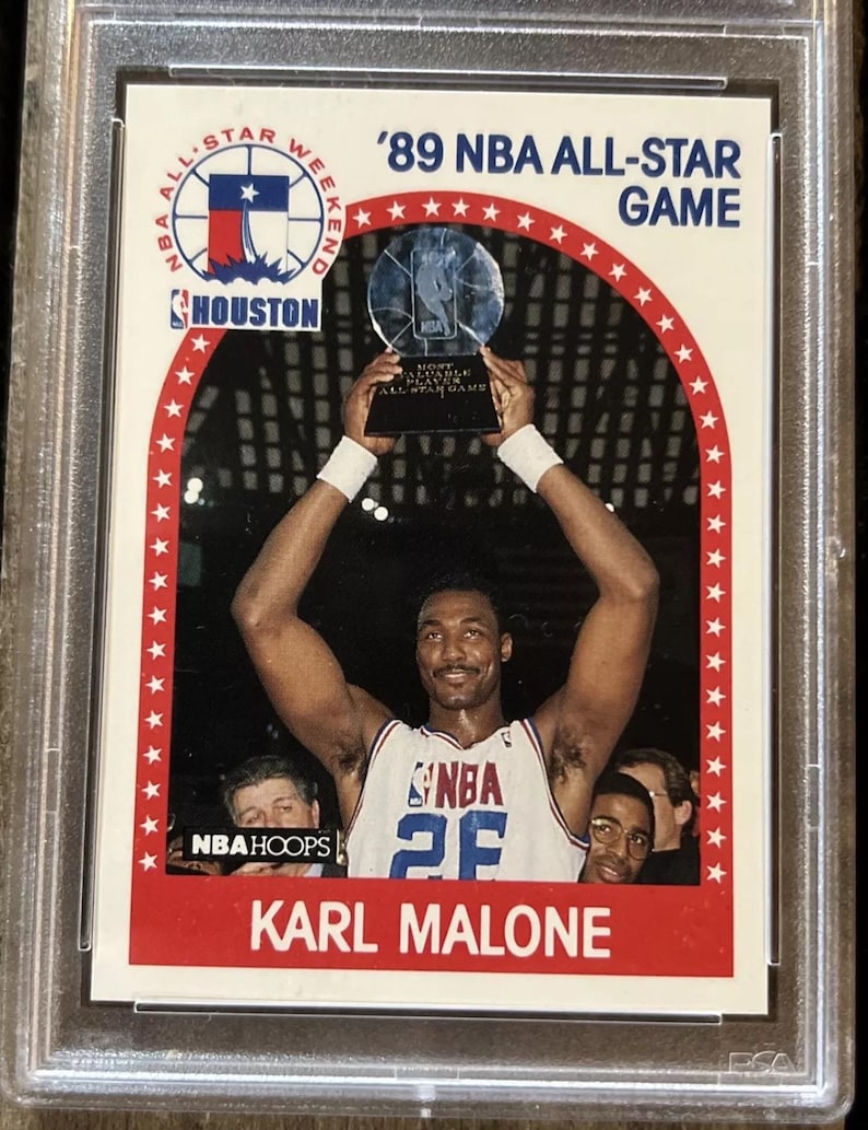 Pu&ograve; includere: Una carta da collezione del NBA All-Star Game con Karl Malone che tiene un trofeo. La carta ha un bordo bianco con uno sfondo rosso e il testo '89 NBA All-Star Game' in bianco.
