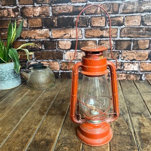 Puede incluir: Una lámpara de aceite de metal roja con un globo de vidrio y un asa de alambre. La lámpara está sobre una superficie de madera.