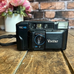 Puede incluir: Una cámara de película Vivitar TEC45 negra de 35 mm con flash incorporado. La cámara tiene una etiqueta azul y blanca que dice "Auto Focus 35mm 1:3.2".