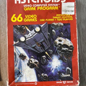 Puede incluir: Una caja roja de cartucho de juego Atari con una nave espacial y asteroides en la parte delantera. La caja está etiquetada como "Asteroids" y "66 Video Games".