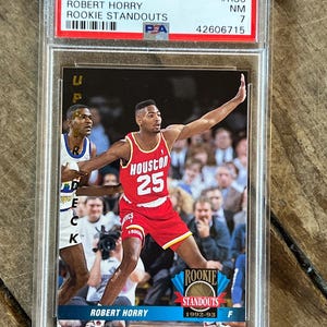 Op de afbeelding: Een 1992 Upper Deck Robert Horry Rookie Standouts basketbalkaart, in een beschermende houder. De kaart toont een speler in een rood Houston Rockets-uniform, nummer 25, met de tekst "ROOKIE STANDOUTS" en "1992-93".