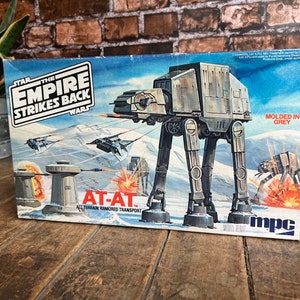 Puede incluir: Una caja para un kit de maquetas de Star Wars del AT-AT, un vehículo de transporte blindado todo terreno. La caja presenta una ilustración colorida del AT-AT caminando sobre un paisaje nevado, con cazas TIE volando por encima. La caja está etiquetada como "Star Wars: El Imperio Contraataca" y "AT-AT: Transporte blindado todo terreno". La caja está hecha por MPC.
