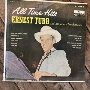 Peut inclure: Pochette de l'album vinyle vintage "All Time Hits" d'Ernest Tubb and his Texas Troubadours. La couverture présente un portrait de Tubb portant un chapeau blanc, tenant une guitare. Le titre de l'album et le nom de l'artiste sont en gros caractères gras.