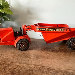 May include: Red toy dump truck with a raised bed and a working arm. The truck is labeled "Lumar Rocker Dump".