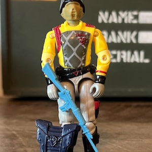 May include: A G.I. Joe action figure wearing a black helmet, a yellow long-sleeve shirt with a grey diamond-patterned chest plate, and light grey trousers. The figure holds a light blue rifle and stands next to a dark blue rectangular shield.