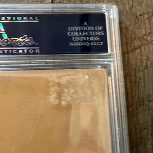 May include: A close-up of a graded sports card in a protective case. The case is clear plastic with a gold border. The card is a light brown color and has a slight sheen. The text on the case reads "A division of Collectors Universe NASDAQ: CLCT".