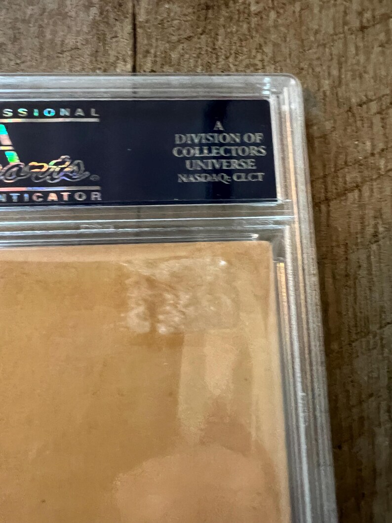 May include: A close-up of a graded sports card in a protective case. The case is clear plastic with a gold border. The card is a light brown color and has a slight sheen. The text on the case reads "A division of Collectors Universe NASDAQ: CLCT".
