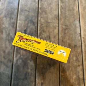 May include: A yellow VHS tape box for "The Adventures of Young Indiana Jones: Treasure of the Peacock's Eye." The box features the Paramount and Lucasfilm logos, with text indicating the chapter number, color, and runtime of 94 minutes.