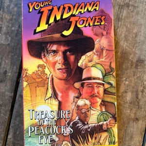 May include: A VHS tape for "The Adventures of Young Indiana Jones: Treasure of the Peacock's Eye." The cover features a portrait of a man in a fedora, with the title in yellow and orange. The background is purple and orange.