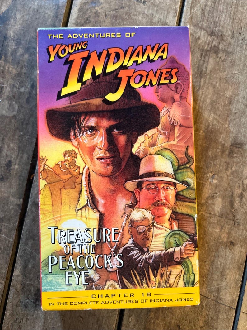 May include: A VHS tape for "The Adventures of Young Indiana Jones: Treasure of the Peacock's Eye." The cover features a portrait of a man in a fedora, with the title in yellow and orange. The background is purple and orange.