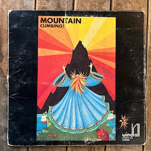 May include: A square vinyl record jacket titled Mountain Climbing! featuring a central illustration of a figure in a blue robe with long orange hair reaching toward a black mountain peak against a yellow and red sunburst background.
