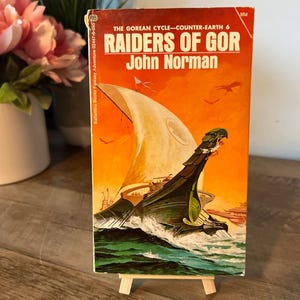May include: A vintage paperback book titled "RAIDERS OF GOR" by John Norman. The cover features a ship sailing on a green sea with an orange sky. The book is propped up on a small wooden easel.