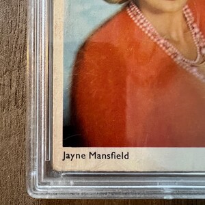 May include: A vintage photograph of Jayne Mansfield, a famous actress, in a red dress with a pearl necklace. The photo is in a plastic protective case.
