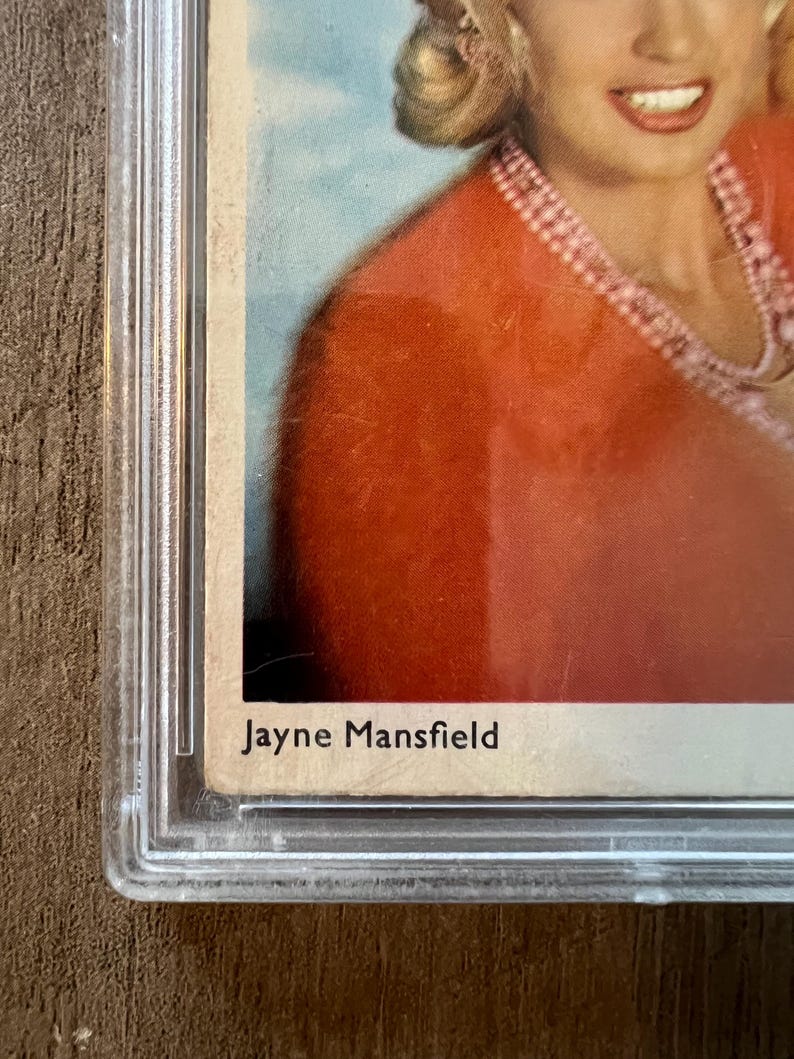 May include: A vintage photograph of Jayne Mansfield, a famous actress, in a red dress with a pearl necklace. The photo is in a plastic protective case.