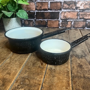 May include: Two black enamel saucepans with white interiors and long handles. The pans have a speckled design and are set on a rustic wooden surface. The background includes a brick wall and greenery.