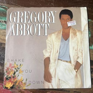 Könnte beinhalten: Albumcover für Gregory Abbotts "Shake You Down". Das Cover zeigt einen Mann in einem cremefarbenen Anzug mit goldenen Akzenten. Der Albumtitel und der Name des Künstlers sind in großen, fetten Buchstaben gedruckt. Ein Aufkleber mit einem Preisschild ist sichtbar.