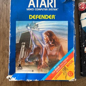 May include: A blue Atari video game box with a picture of a woman and a spaceship on the front. The game is called "Defender" and the box includes a comic book.
