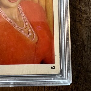 May include: A close-up of a vintage photograph of a woman wearing a red sweater and a pearl necklace. The photo is framed and has the number "63" written on the bottom right corner.