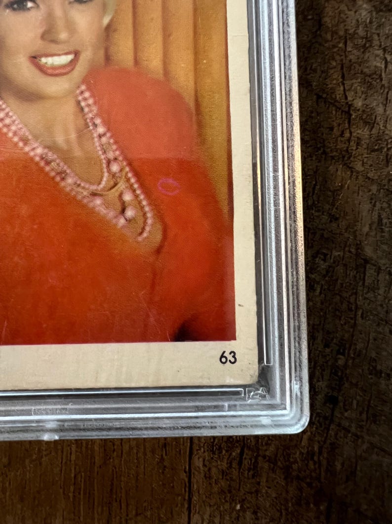 May include: A close-up of a vintage photograph of a woman wearing a red sweater and a pearl necklace. The photo is framed and has the number "63" written on the bottom right corner.